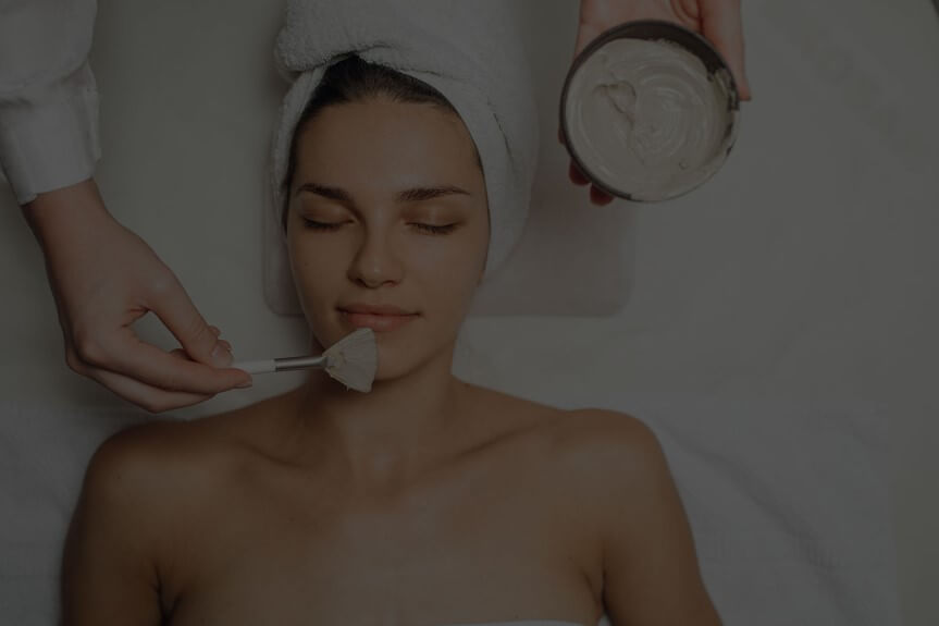 Person lying down with eyes closed, a towel wrapped around their head, receiving a facial treatment 